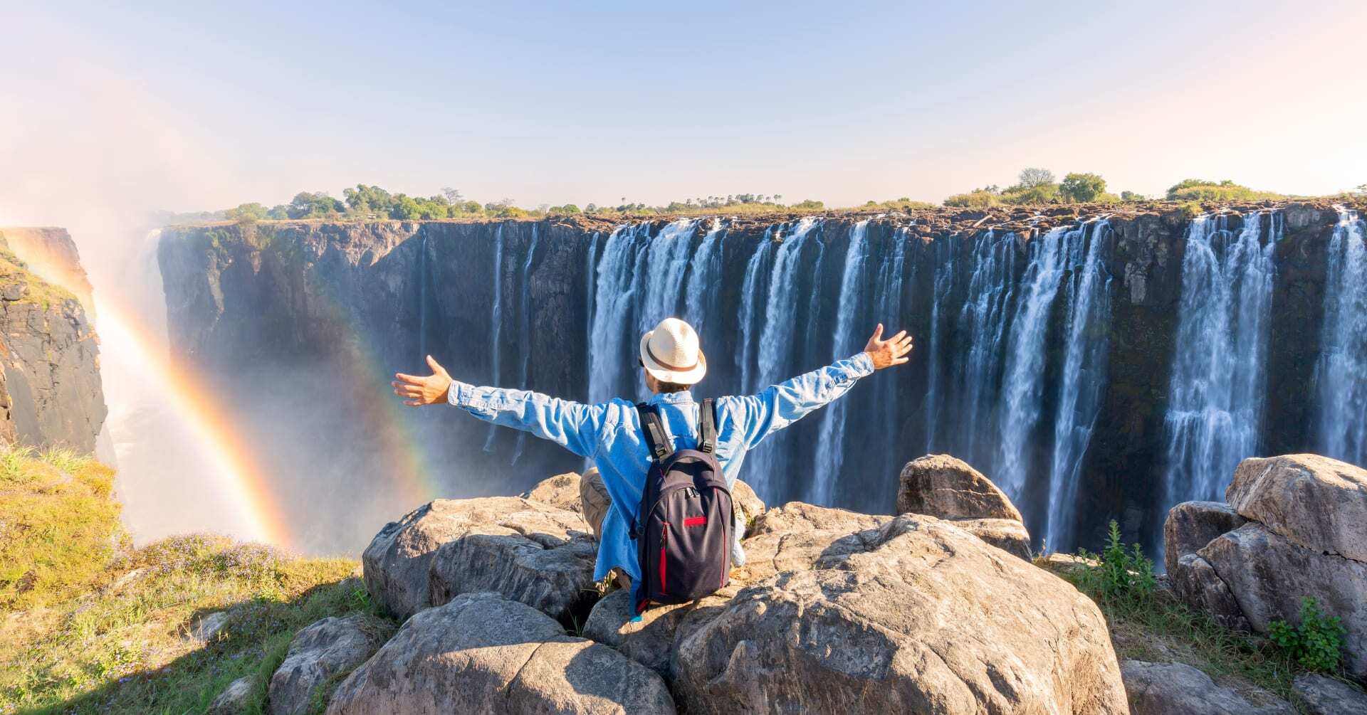 A visitor sitting on the rocks looking out over Victoria Falls as a rainbow forms in the spray