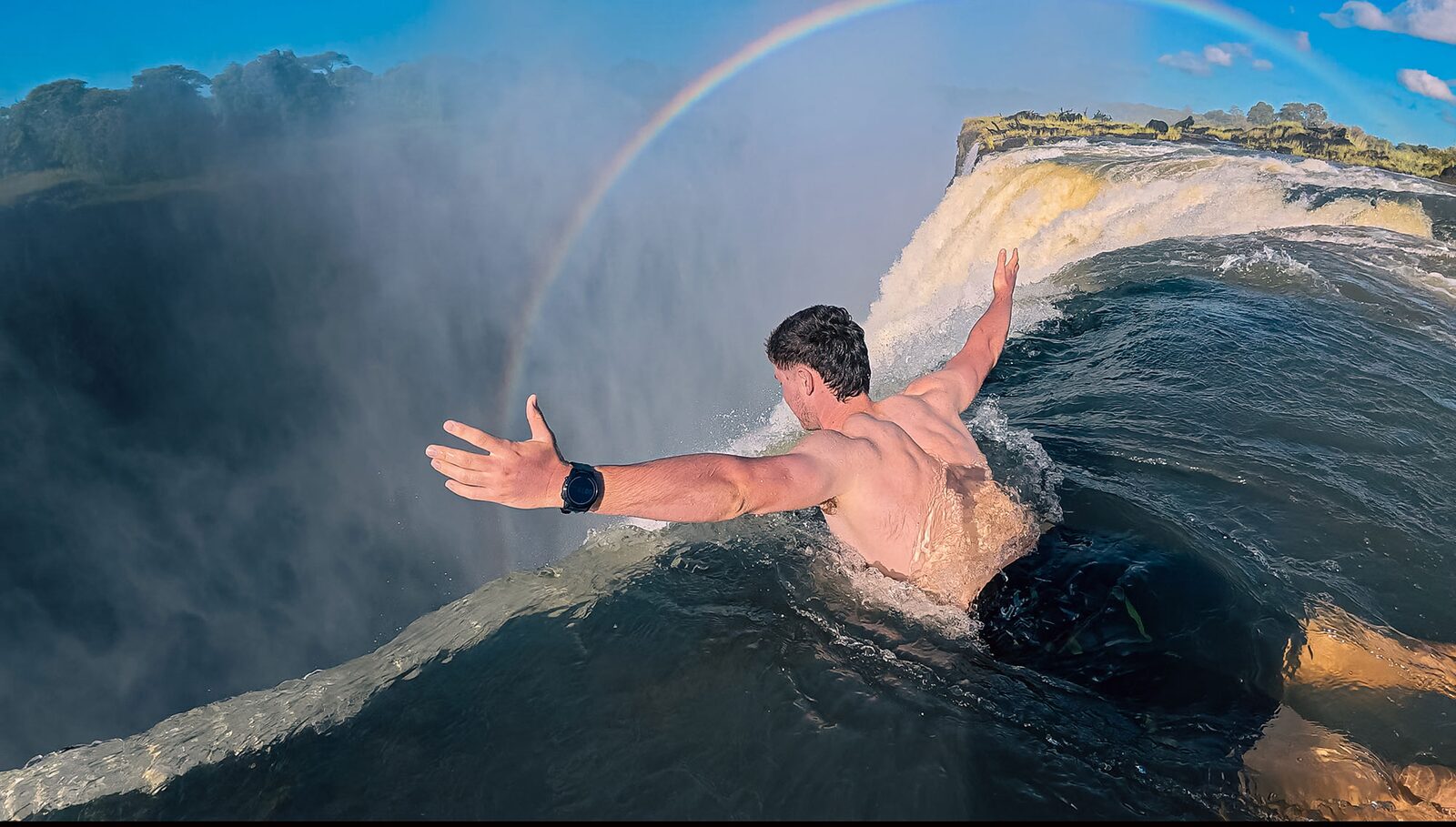 A swimmer at the edge of Devil’s Pool on the lip of Victoria Falls on the Zambian side, with a rainbow behind